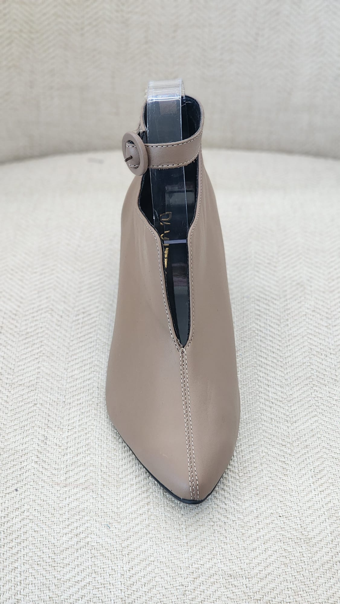 Vista frontal de zapato beige con punta afilada y detalle de correa al tobillo con hebilla redonda.