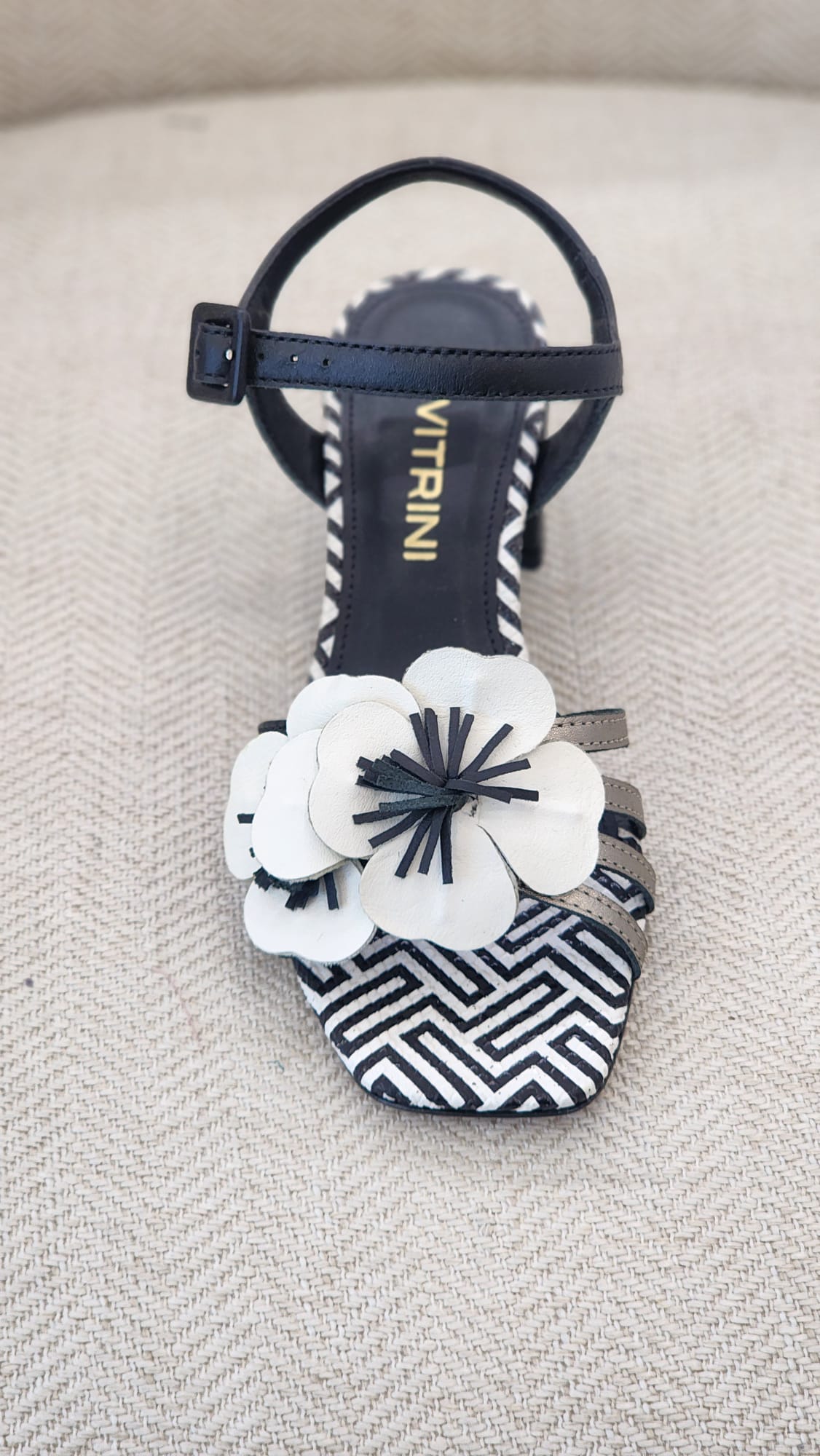 Vista frontal de sandalia preta con correa al tobillo, flor decorativa y punta cuadrada con estampado geométrico.